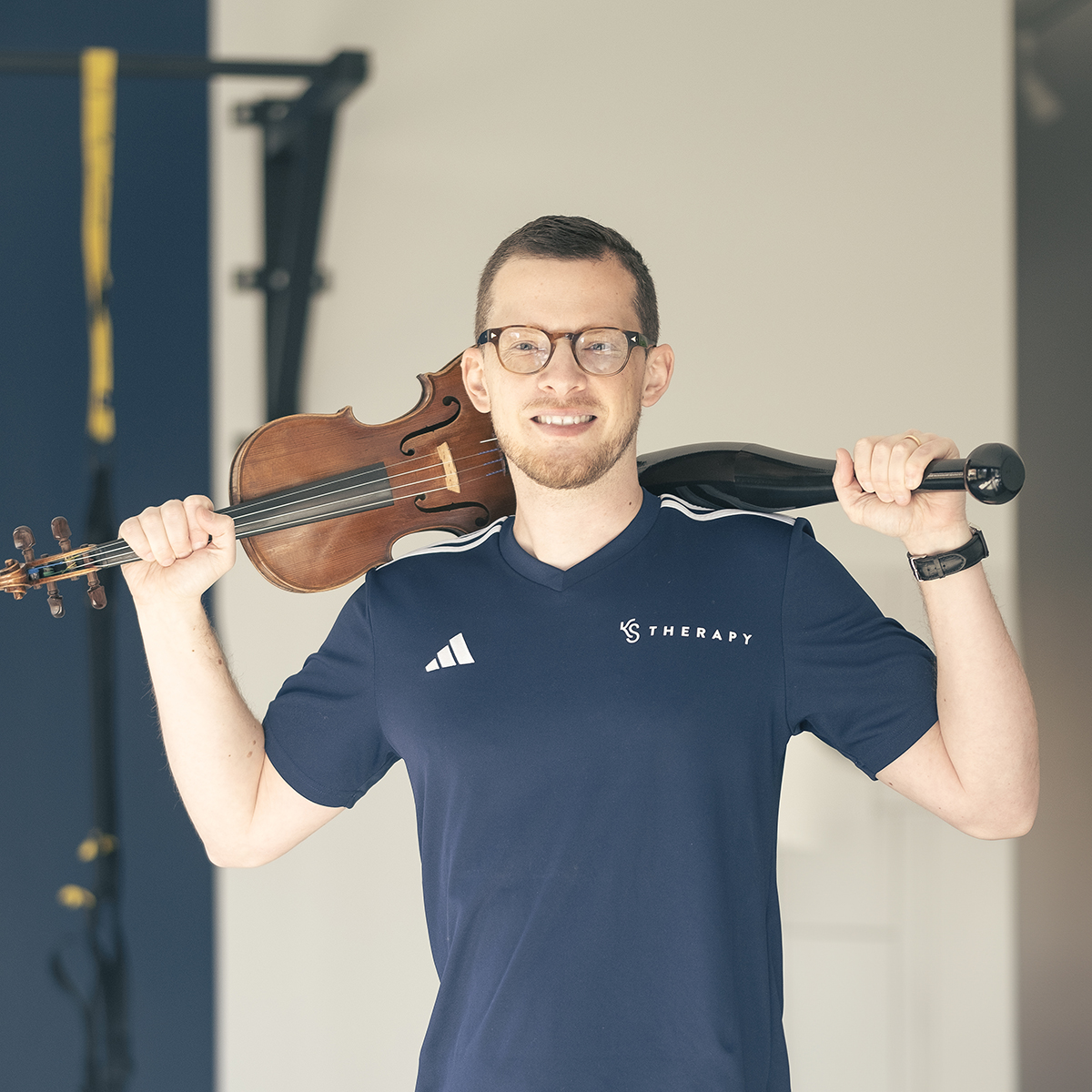 Portrait d'un Physiothérapeute des Musicien à Genève dans une salle de sport. Il tient un violon sur son épaule droite et un Clubbel sur son épaule Gauche.
